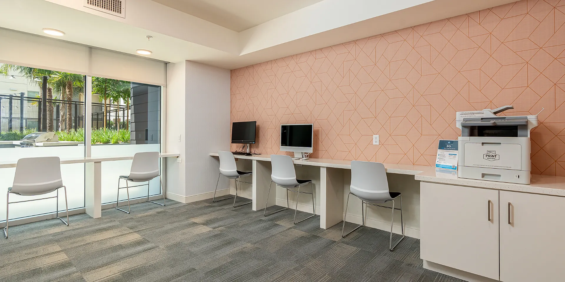 Modern coworking space with sleek computers, stylish seating, and vibrant geometric wall design. Inviting atmosphere with abundant natural light from large windows overlooking lush greenery.