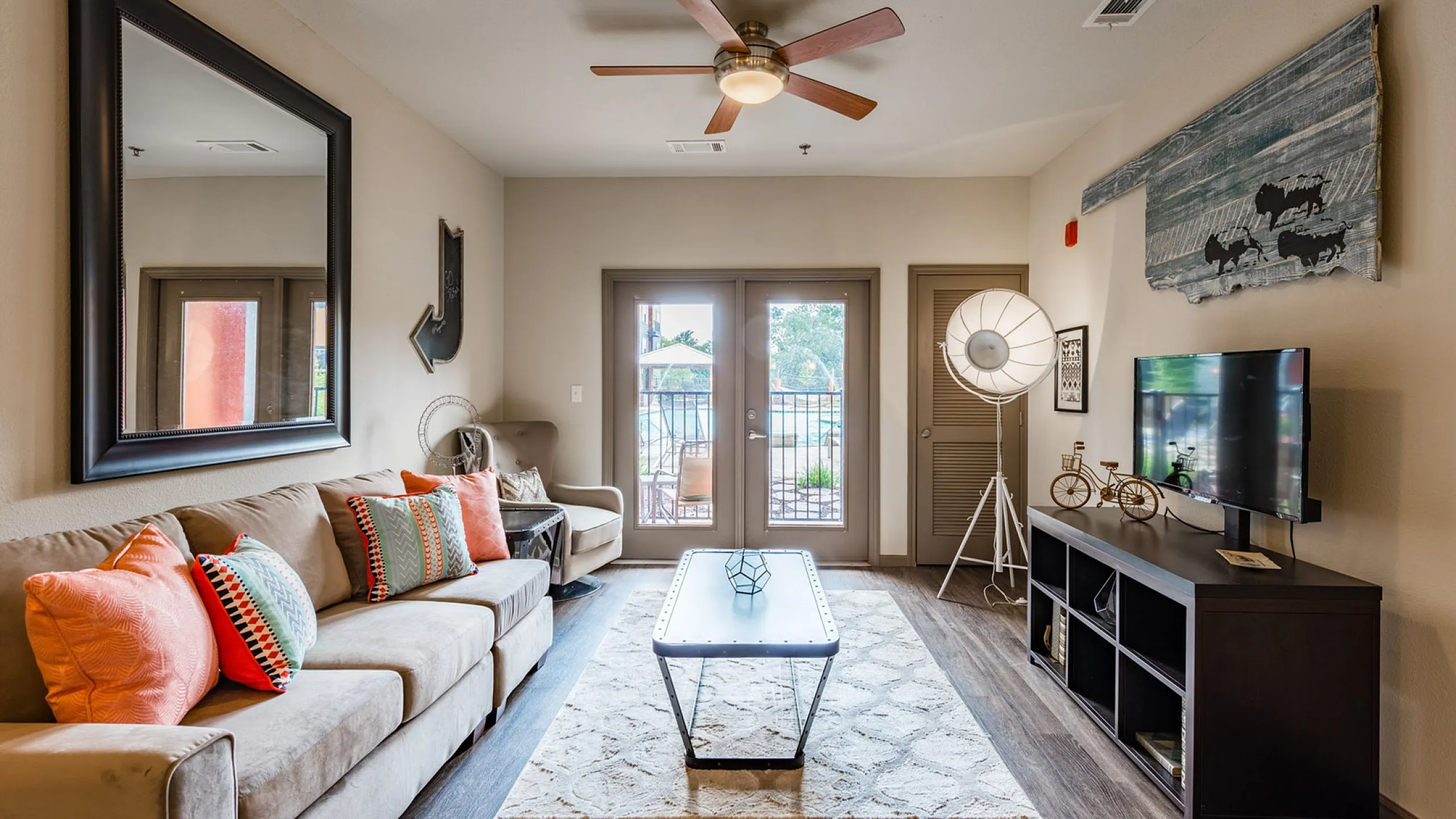 Inviting living room with modern decor, vibrant pillows on a plush sofa, a ceiling fan, and a view of the outdoor pool area through large French doors.
