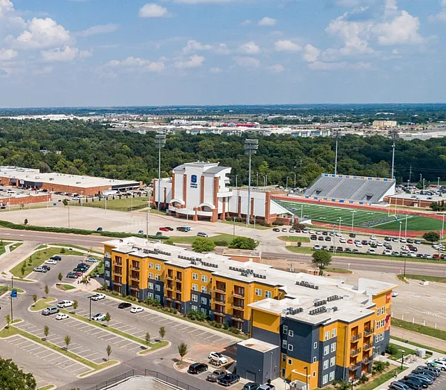 Vibrant aerial view of a modern apartment complex surrounded by lush greenery, with ample parking and stunning lake views in a lively community setting.