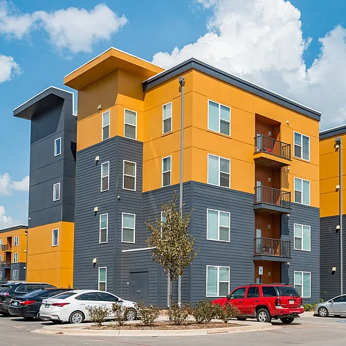 Modern apartment complex with vibrant yellow and gray design, featuring multiple balconies, surrounded by ample parking and lush greenery under a clear blue sky.