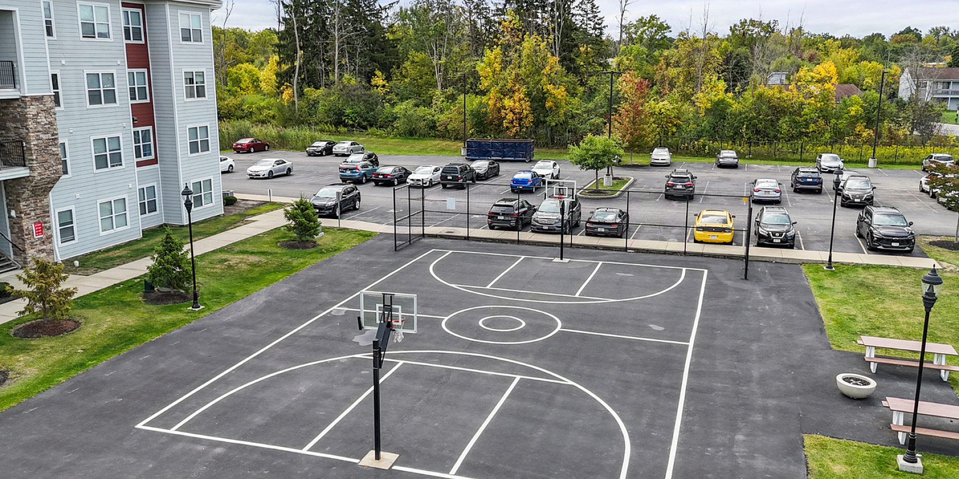 Modern apartment complex with sleek stone accents, adjacent to a spacious parking lot and well-maintained basketball court surrounded by lush greenery.