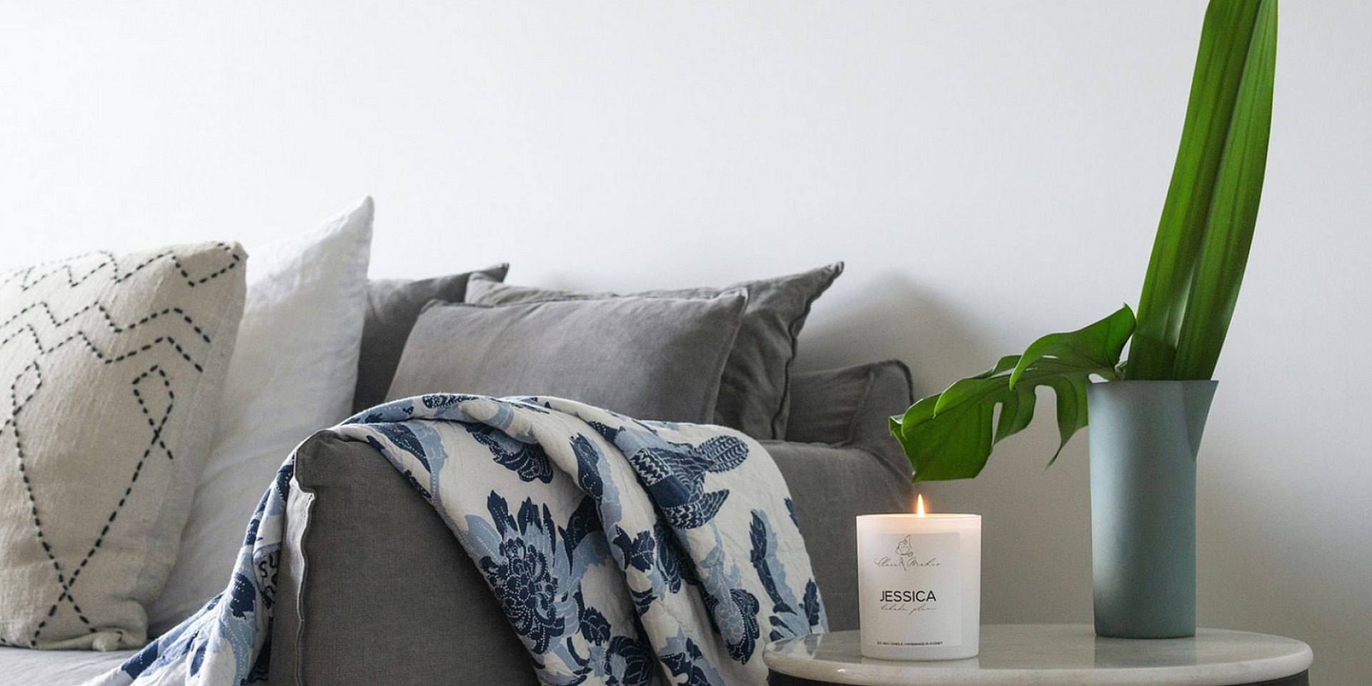 Cozy living area with elegant gray sofa, decorative cushions, and a stylish blue throw blanket. A lit candle and a sleek vase with a green leaf sit on a modern round side table, adding warmth and sophistication.