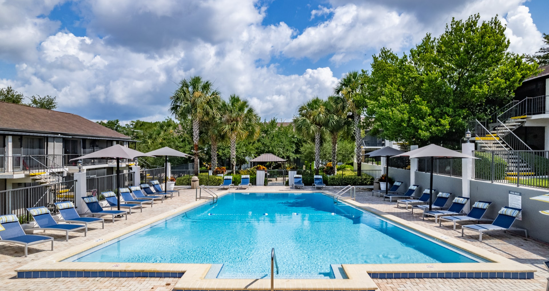 Inviting apartment complex with a sparkling outdoor pool surrounded by lush palm trees and stylish lounge chairs under umbrellas, offering a relaxing oasis.