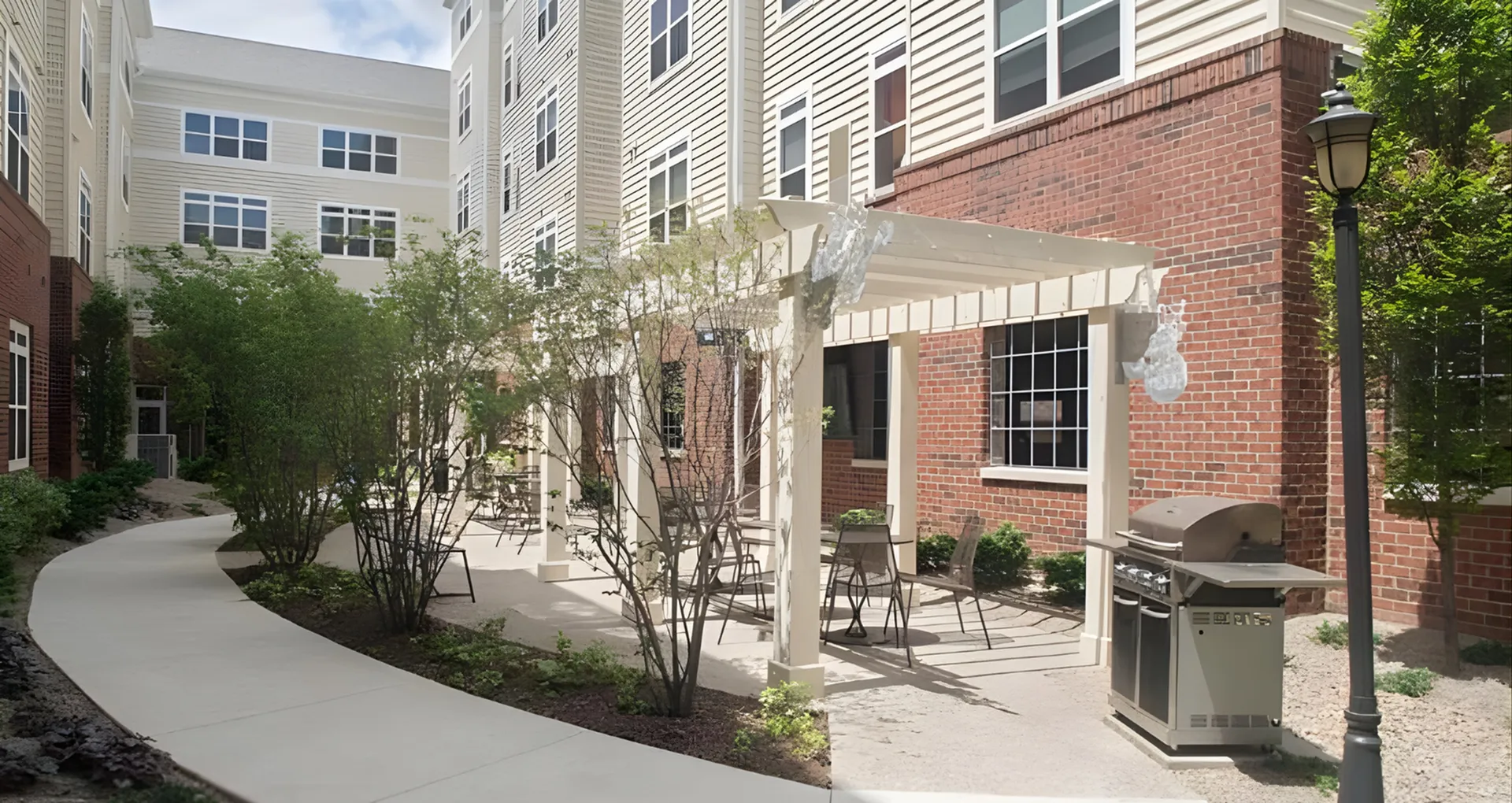 Charming apartment courtyard with a shaded pergola, lush greenery, and modern outdoor grill, perfect for relaxation and gatherings.