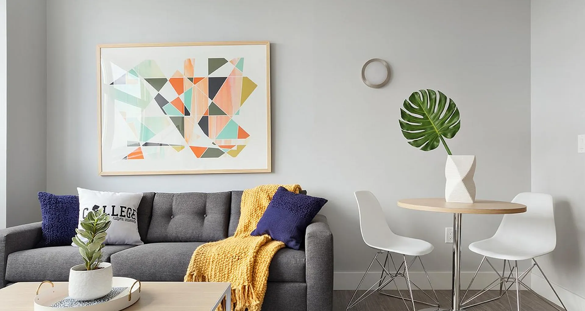 Modern living room with a stylish gray sofa accented by colorful cushions and a cozy yellow throw, complemented by chic geometric wall art and sleek dining nook with white chairs and a leafy plant.