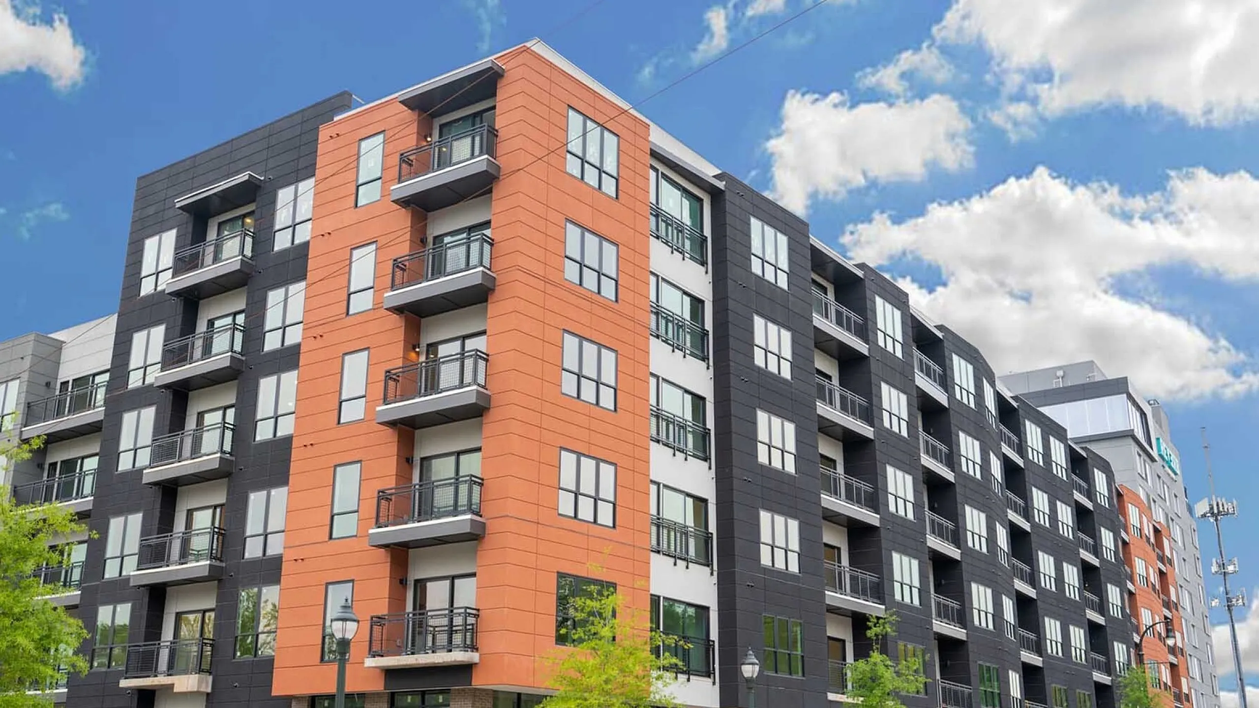Modern urban apartment building with vibrant orange and black facade, featuring private balconies and street-facing views under a bright blue sky.