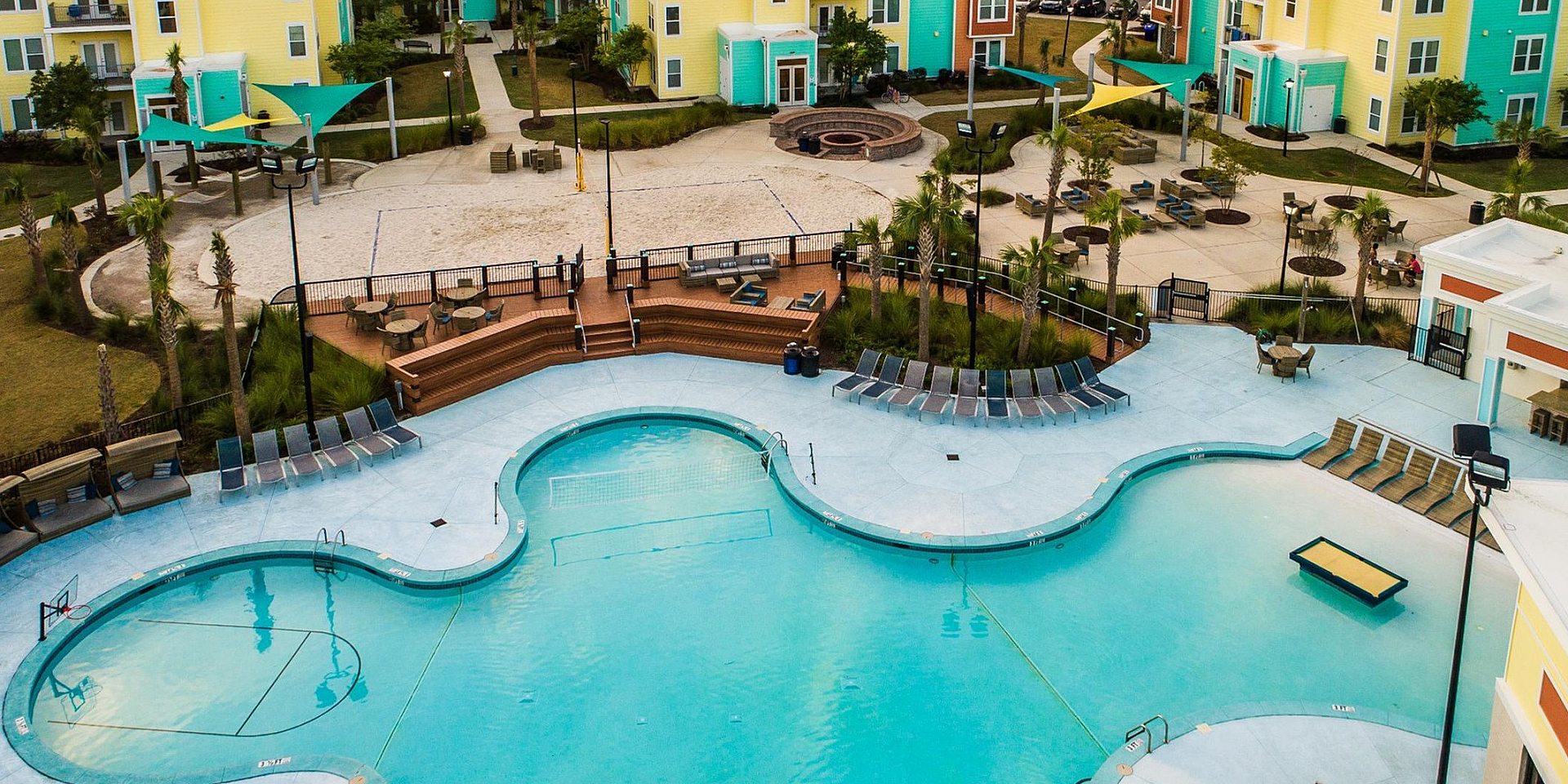 Vibrant apartment complex with colorful buildings, a resort-style pool, and lush palm trees under a stunning sunset sky.