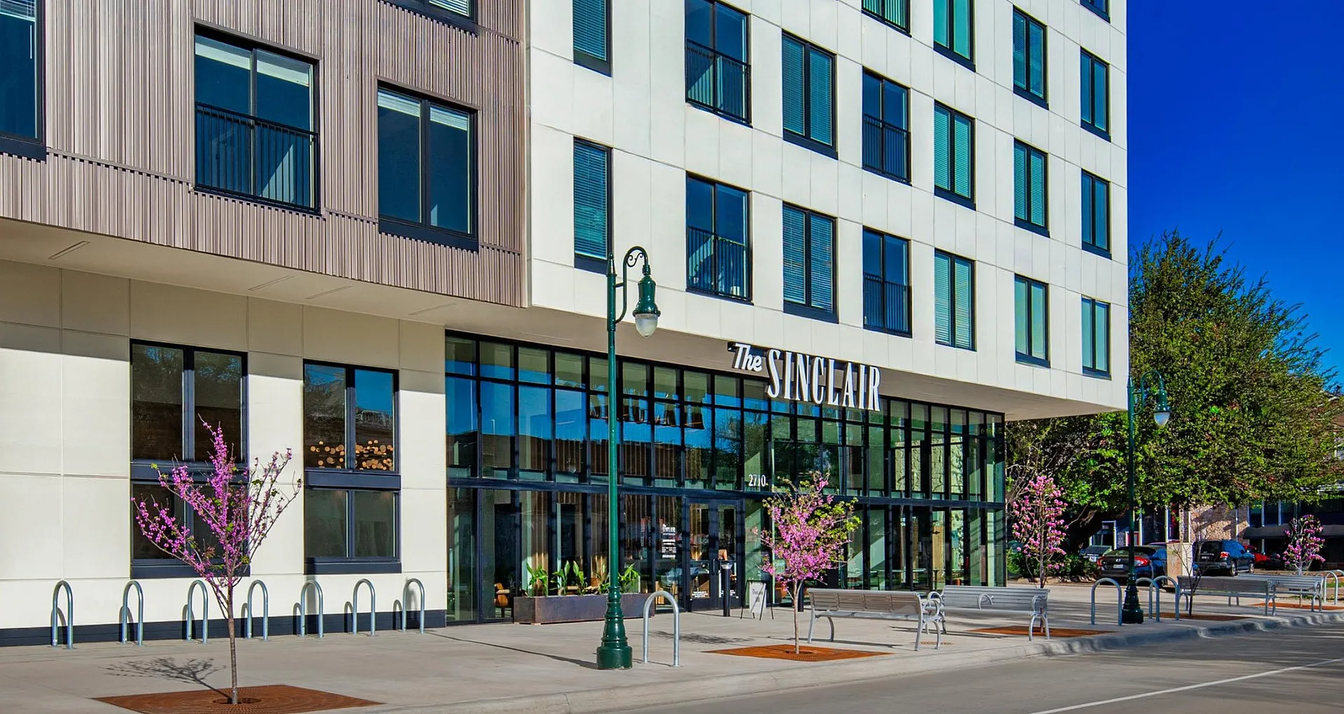 Modern apartment building with sleek design, large windows, and a welcoming entrance surrounded by blooming trees under a clear blue sky.