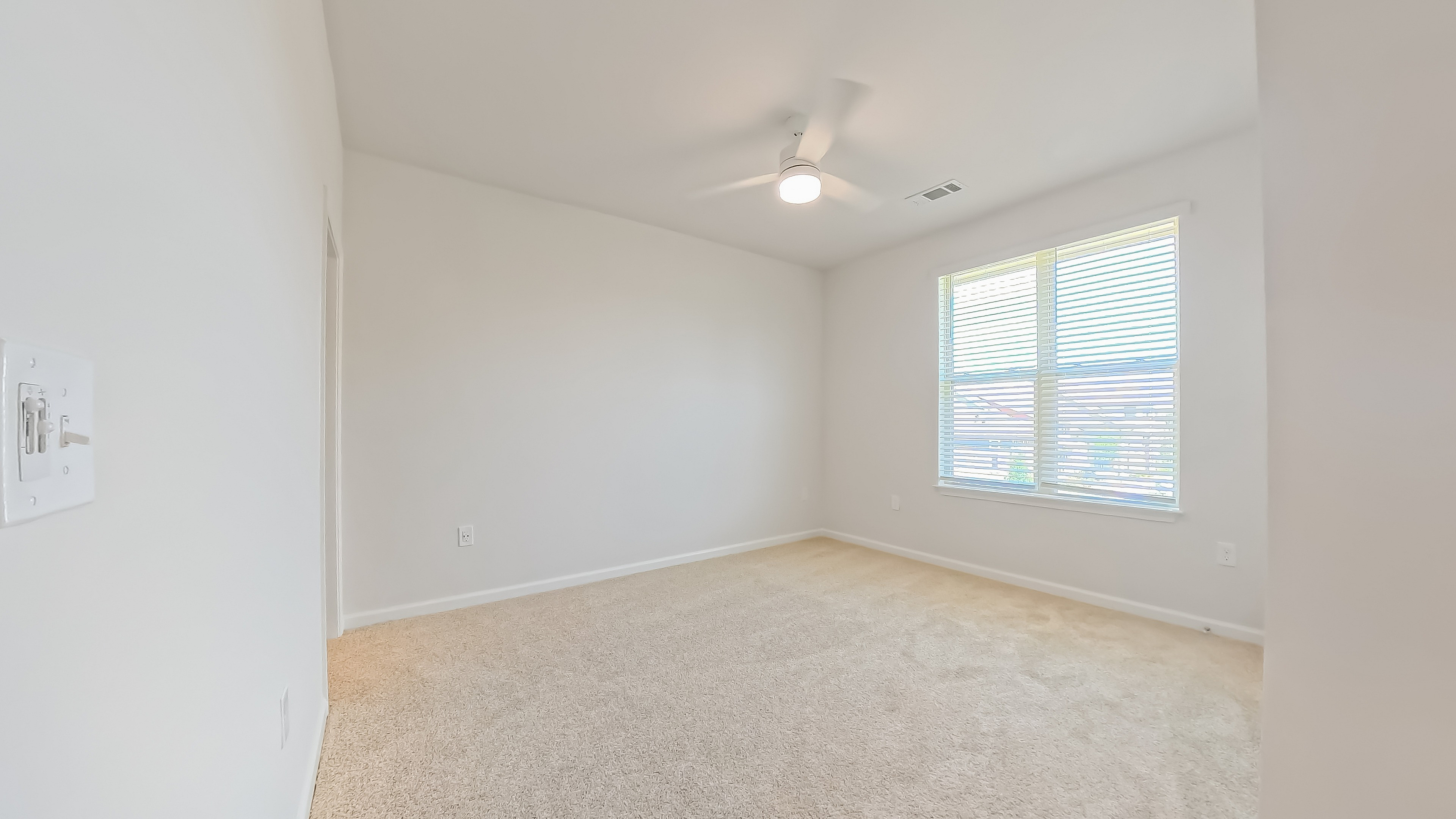 Bright, cozy room with plush carpeting and a ceiling fan, illuminated by natural light from a large window with blinds.