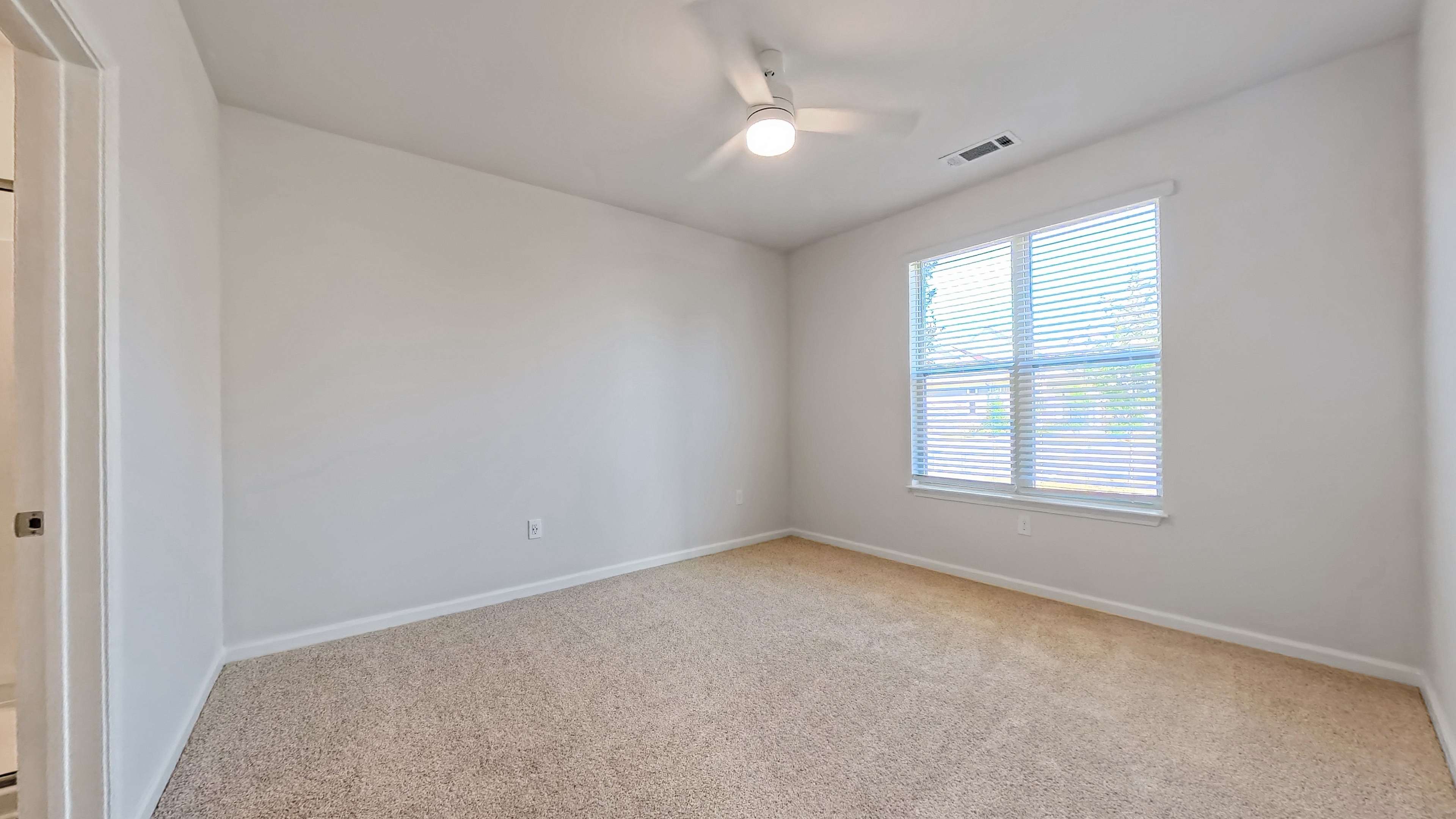 Bright and airy bedroom with plush carpet, large window offering natural light, and a modern ceiling fan.