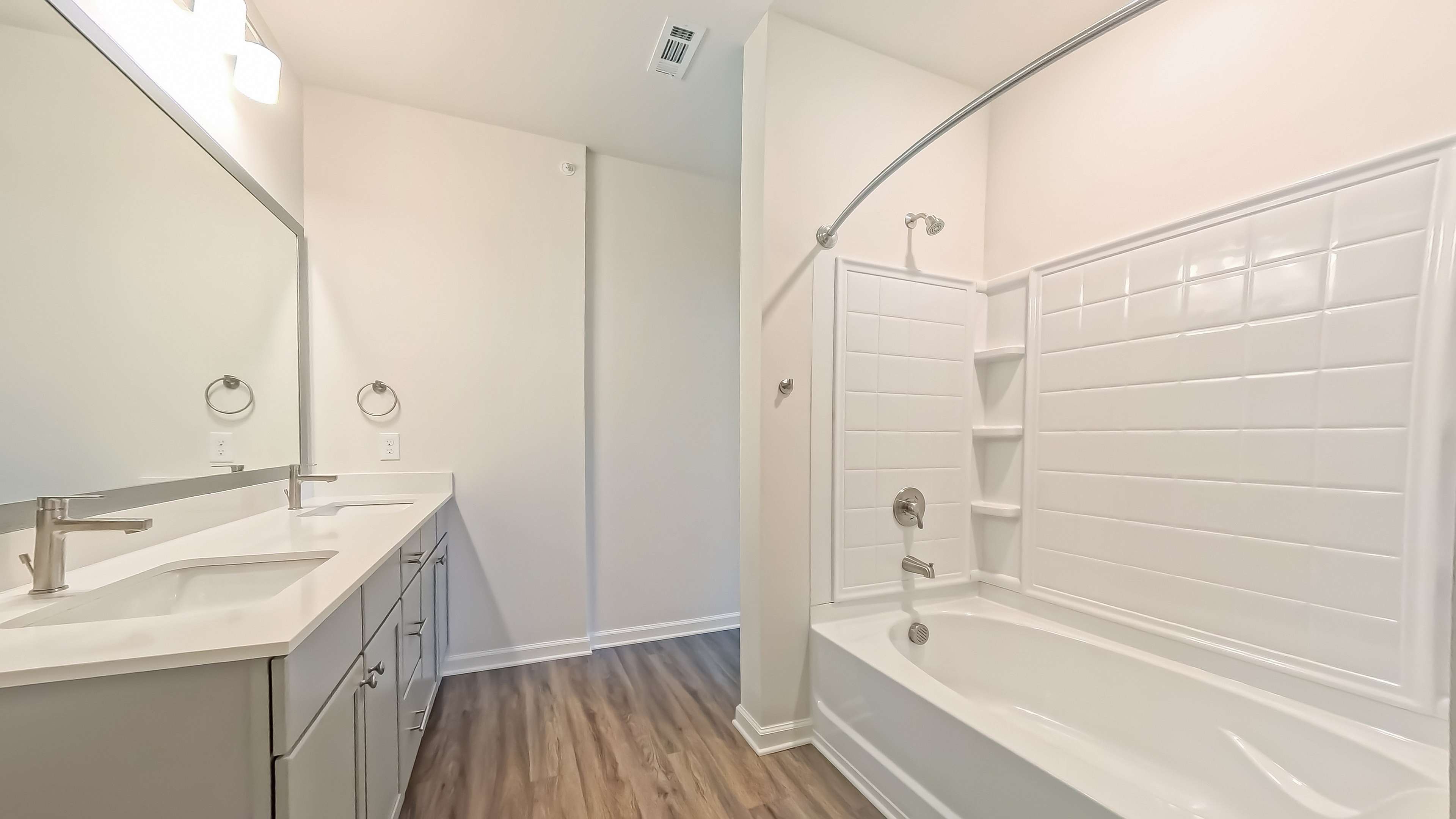 Modern bathroom featuring sleek double sinks, elegant mirror, and a pristine tiled bathtub, complemented by warm wooden flooring.