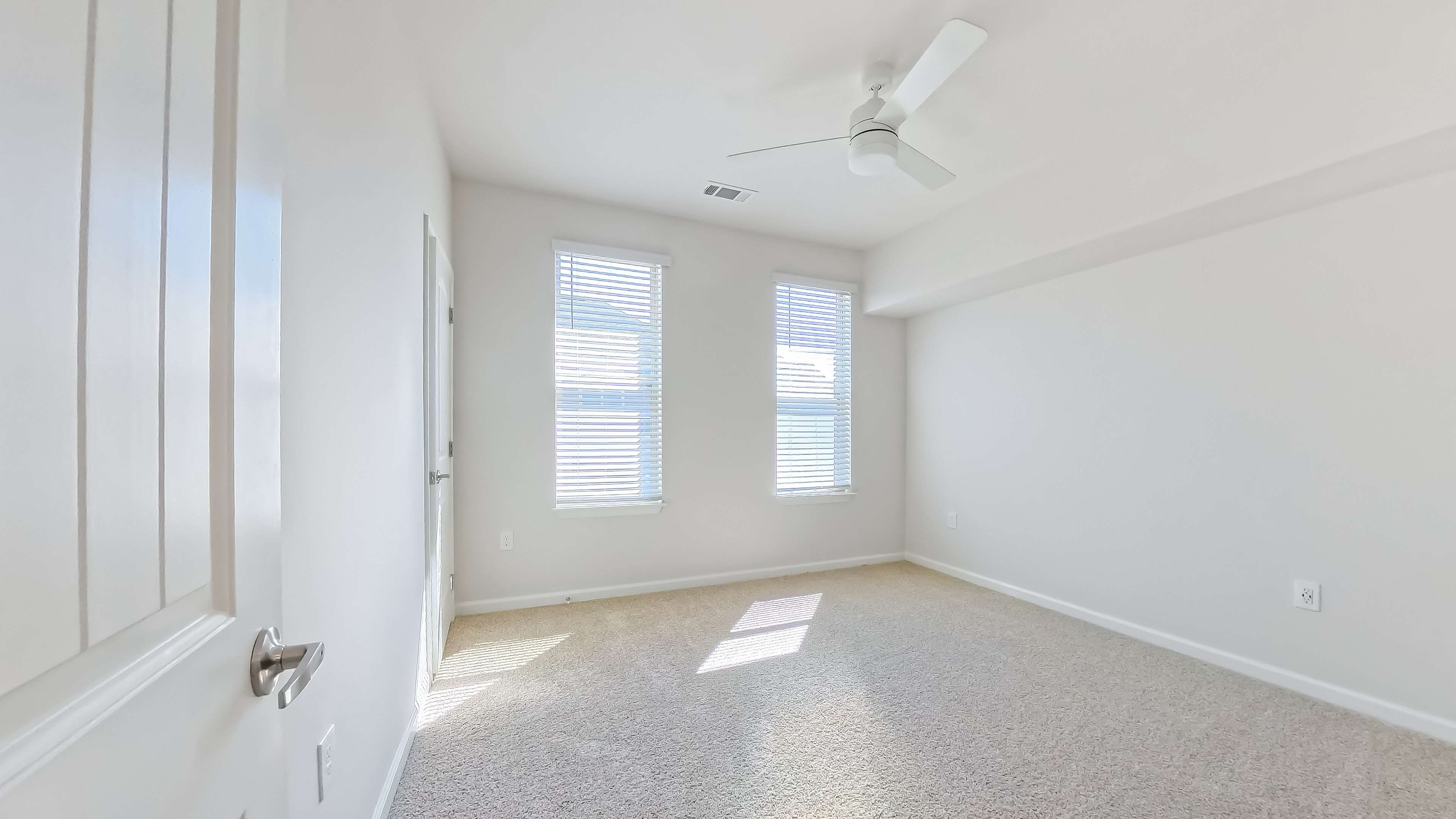 Bright, airy room with plush carpet, two large windows inviting natural light, and a ceiling fan for comfort.