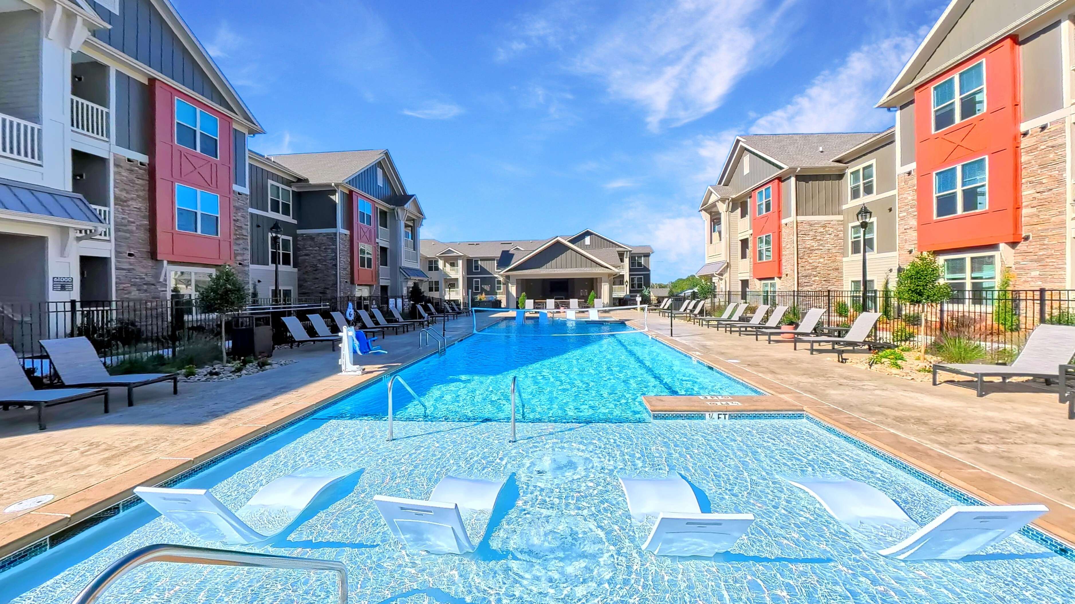 Luxurious apartment complex featuring a stunning outdoor pool with lounge chairs, surrounded by stylish modern buildings and lush landscaping under a clear blue sky.