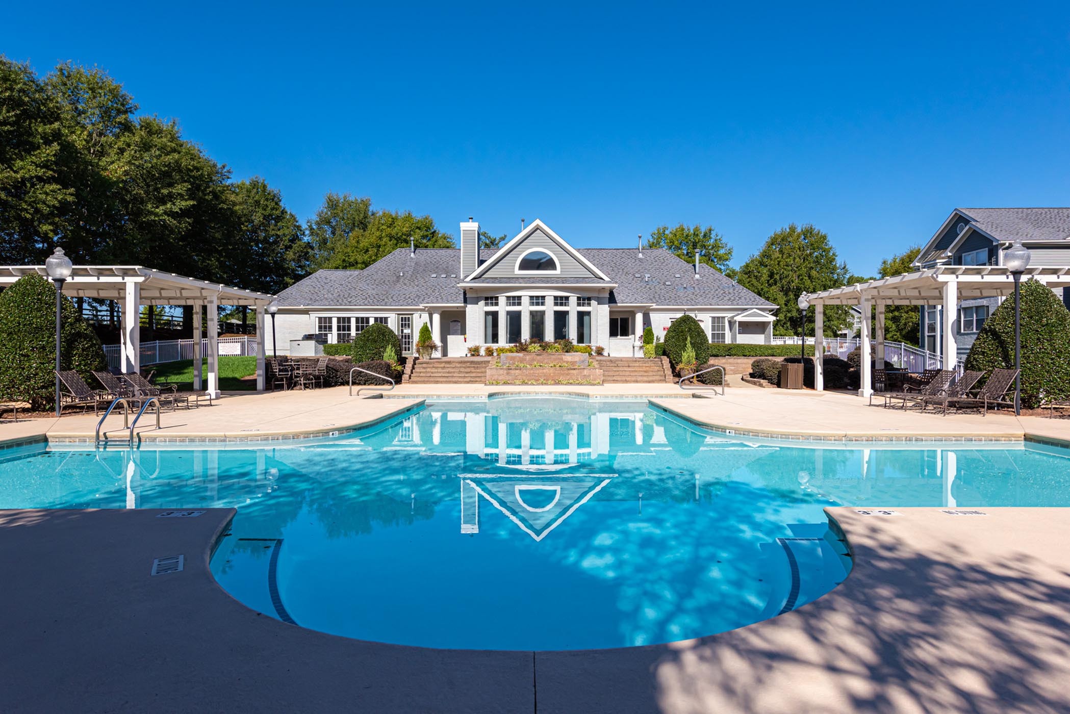 Luxurious apartment complex with a pristine swimming pool, elegant pergolas, and lush landscaping under a clear blue sky.