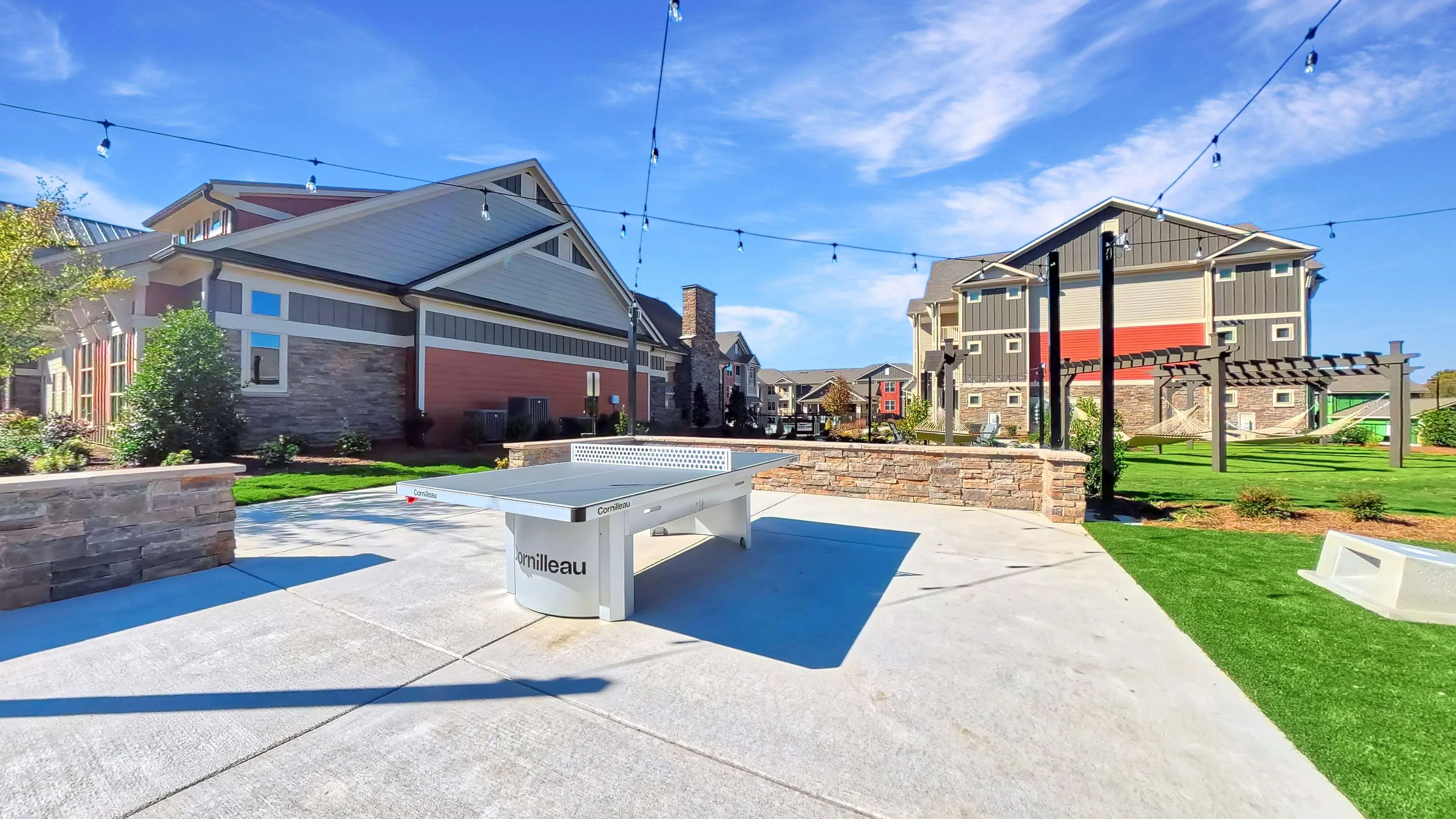 Vibrant apartment complex courtyard featuring a modern ping pong table, lush green landscaping, and ample outdoor seating under a clear blue sky.