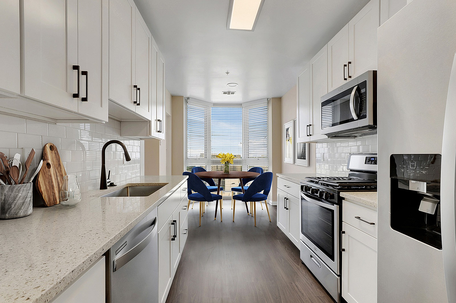 Modern kitchen with sleek white cabinets, stainless steel appliances, and a cozy dining area with blue chairs by large windows.