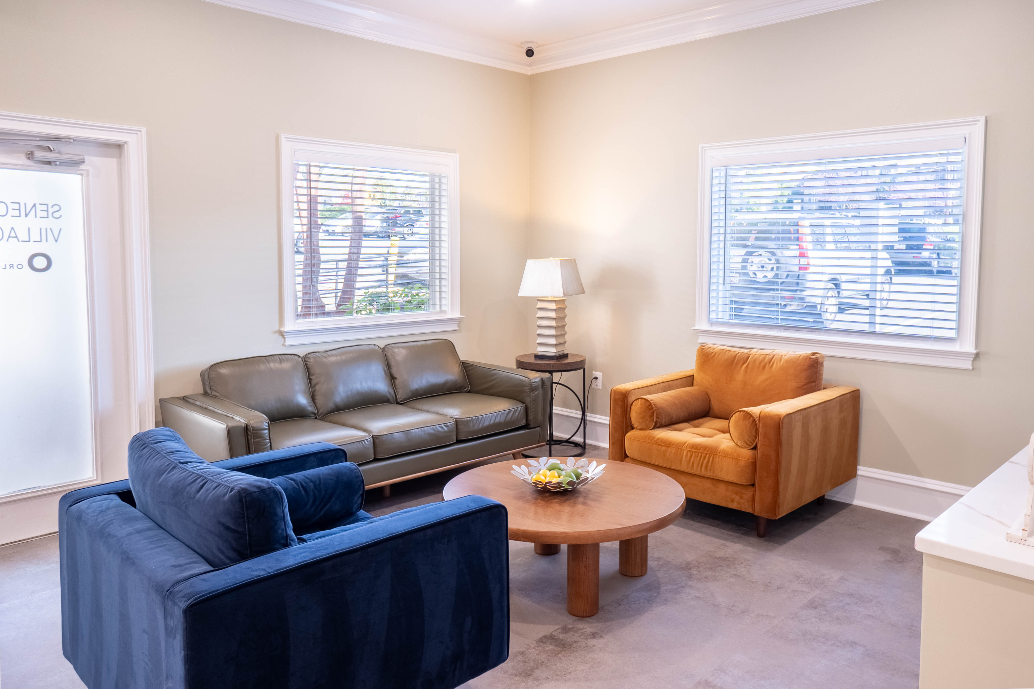 Inviting living room with modern furniture including a plush blue armchair, an orange armchair, a sleek gray sofa, and a stylish round wooden coffee table, bathed in natural light.