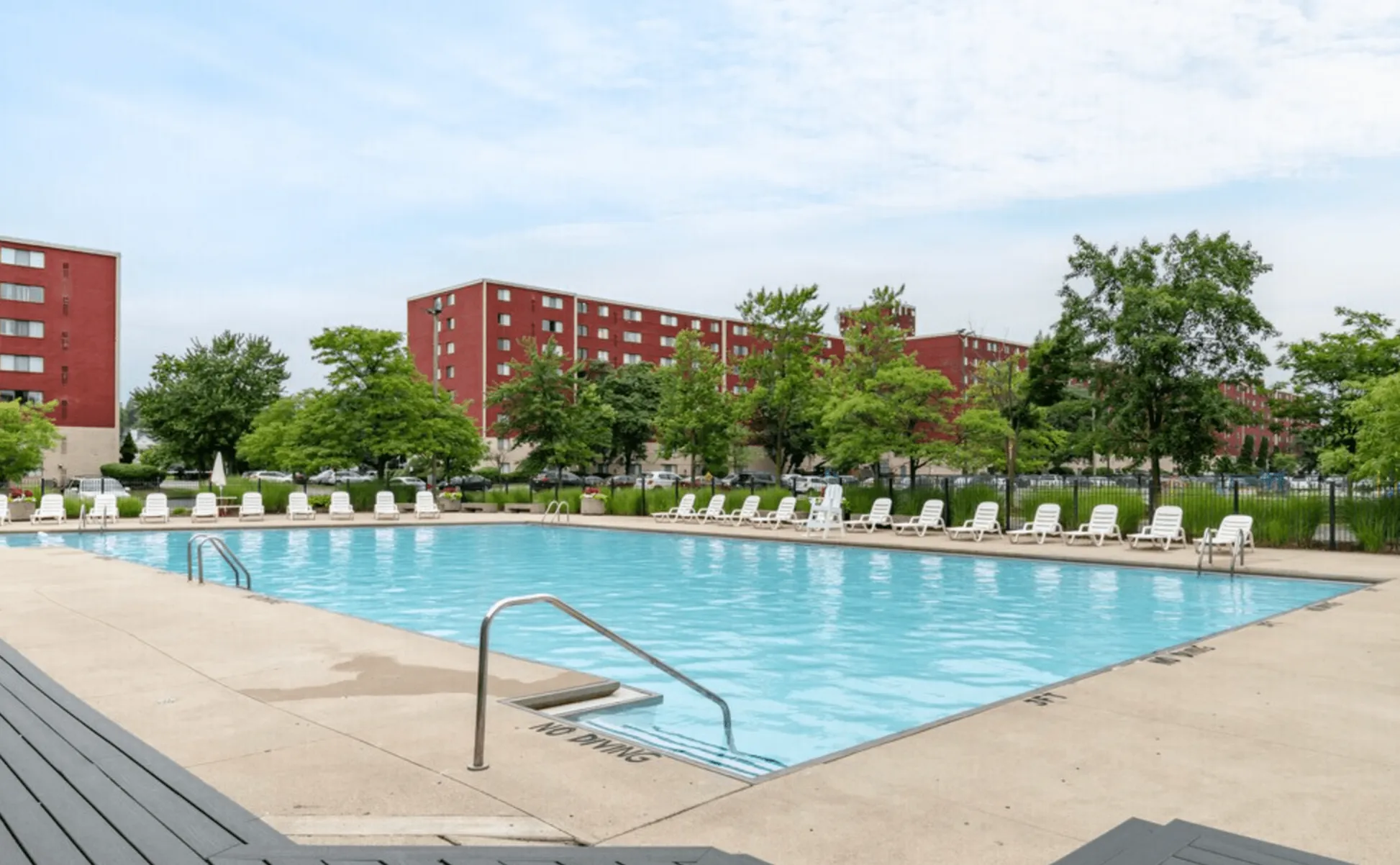 Inviting outdoor pool area with comfortable lounge chairs surrounded by lush greenery and red brick apartment buildings.