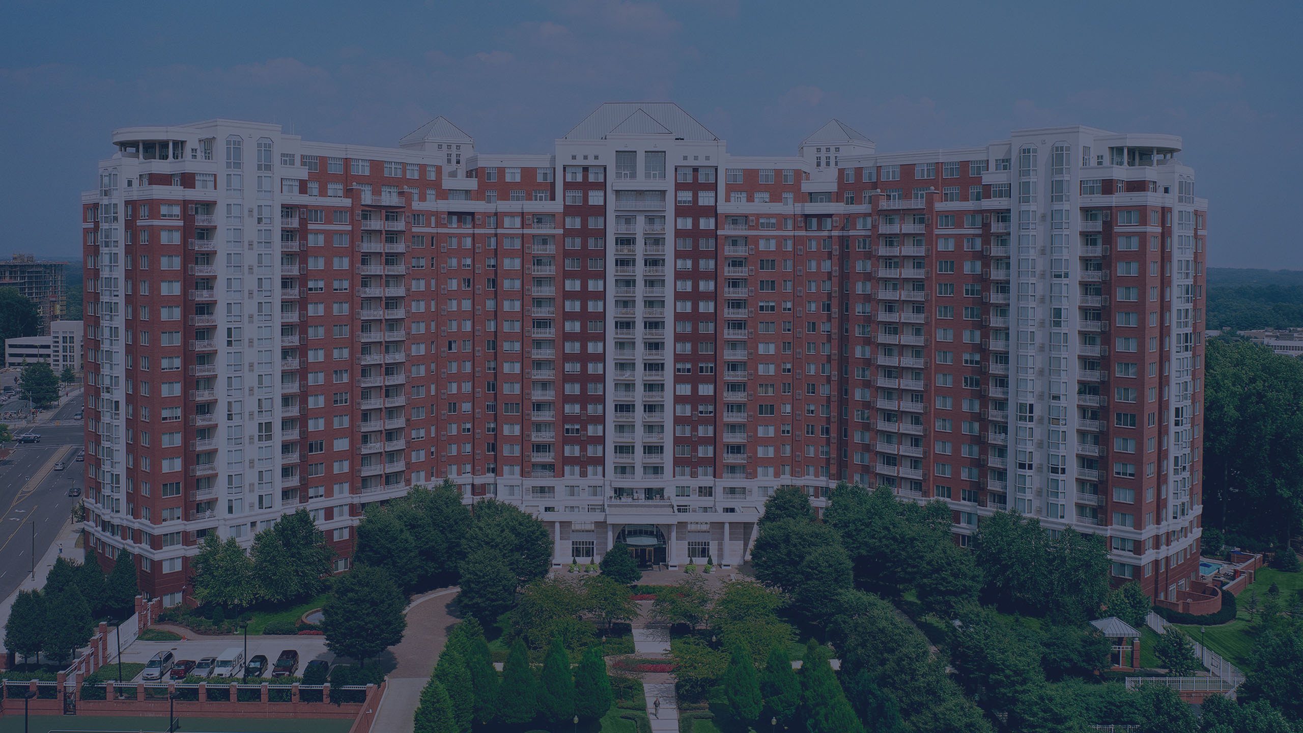 Elegant high-rise apartment building with brick and white facade surrounded by lush greenery and garden pathways, offering urban living with scenic views.