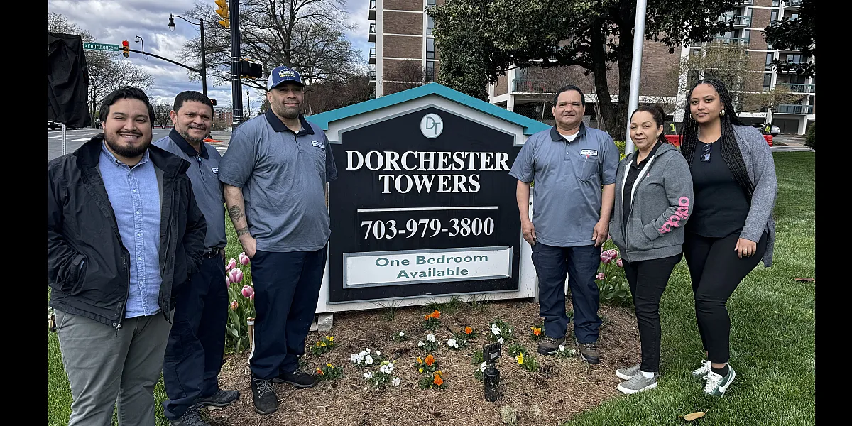 Friendly Dorchester Towers staff standing proudly by the vibrant flower bed and the welcoming sign, advertising one-bedroom availability, showcasing community and quality living.