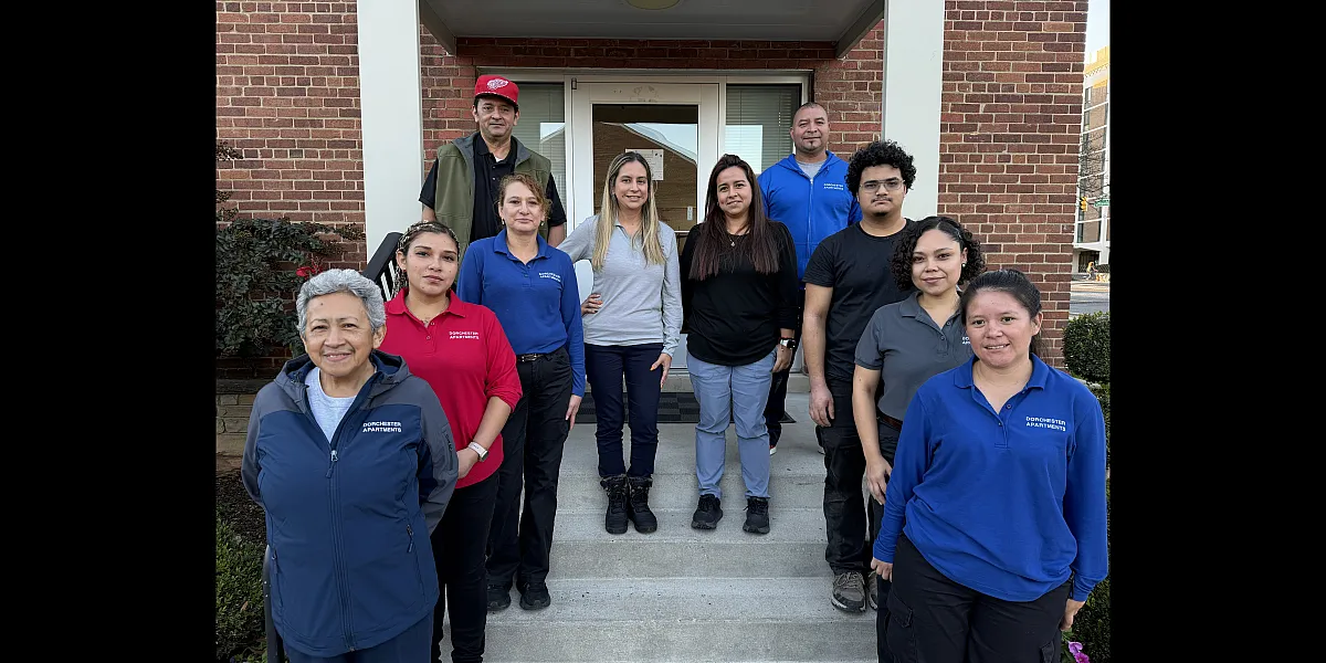 Friendly team of Dorchester Apartments staff standing in front of a welcoming brick entrance, ready to assist residents with a smile.