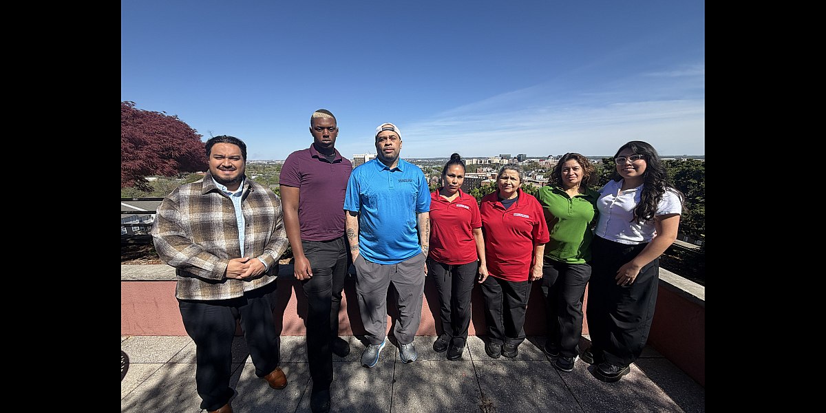 A diverse group of seven people standing on a scenic rooftop terrace with an expansive city skyline view under a clear blue sky.