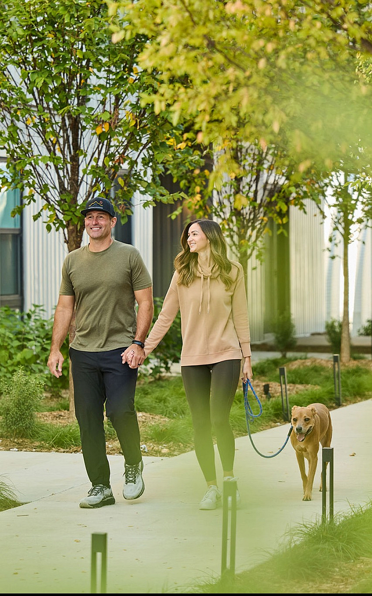 Happy couple enjoys a stroll with their dog through a lush, inviting apartment community path, surrounded by greenery and modern buildings.