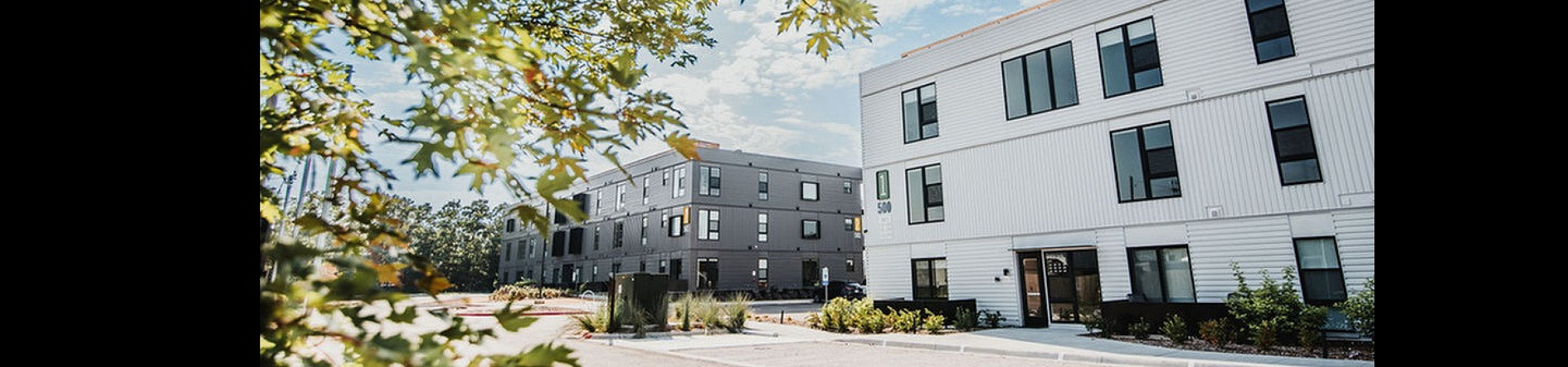 Modern apartment complex with sleek white facade and large windows, surrounded by lush trees under a sunny blue sky.