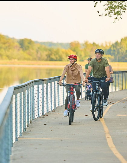 Two people enjoy a scenic bike ride along a lakeside pathway, surrounded by lush greenery and tranquil water reflections.