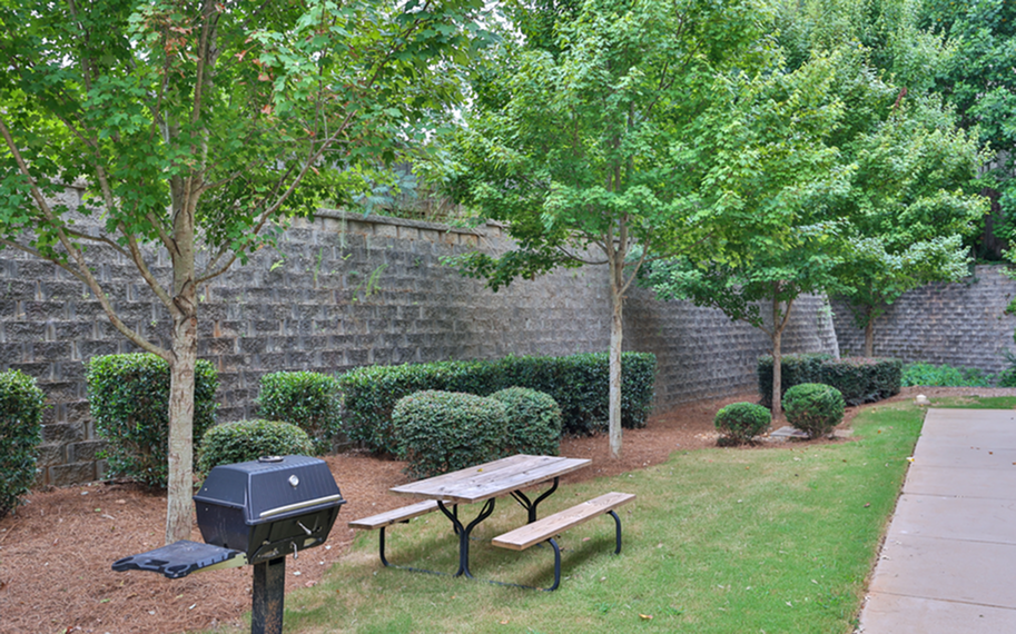 Serene outdoor picnic area with lush greenery, a wooden bench, and a modern grill, perfect for relaxing gatherings.