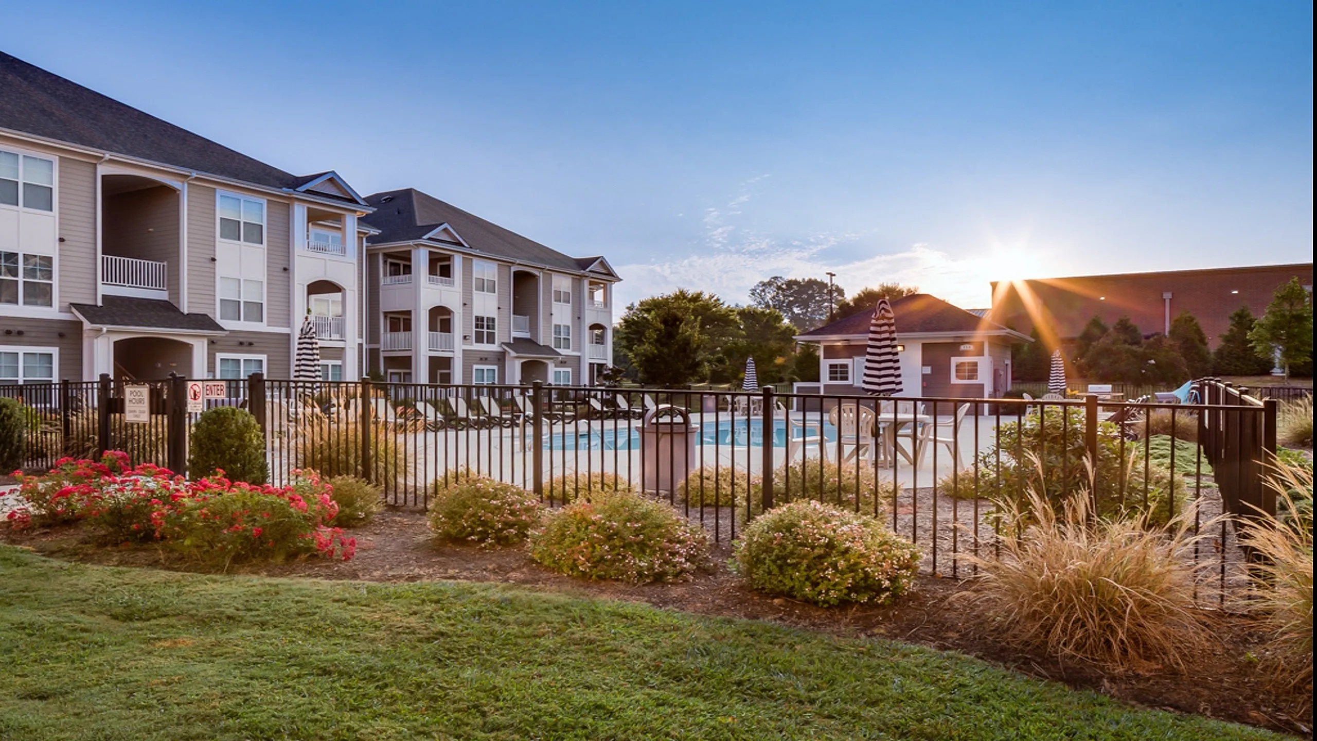 Charming apartment complex featuring modern three-story buildings, an inviting swimming pool, and lush landscaped gardens under a vibrant sunrise.