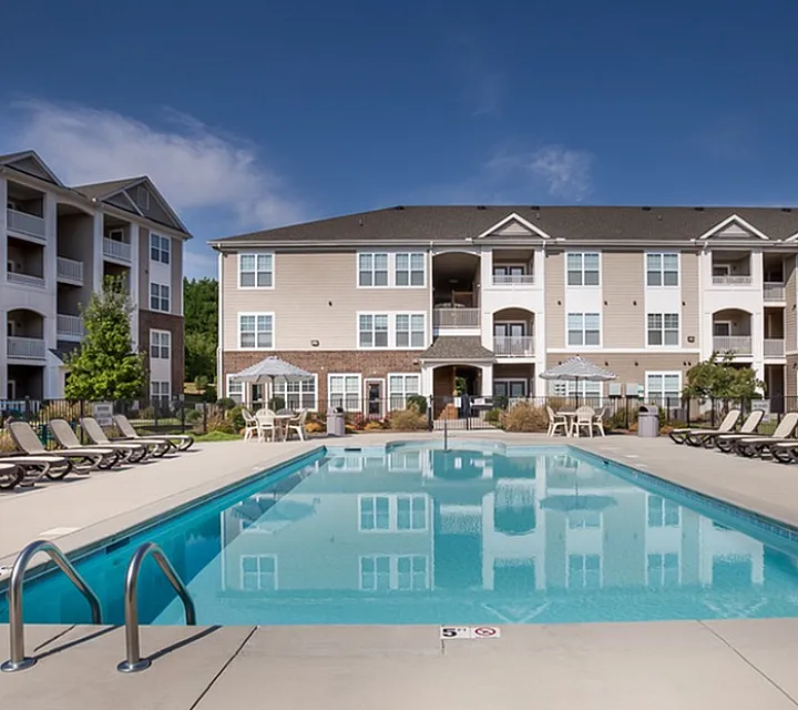 Luxurious apartment complex with resort-style swimming pool, surrounded by sun loungers and elegant multi-story buildings under a clear blue sky.
