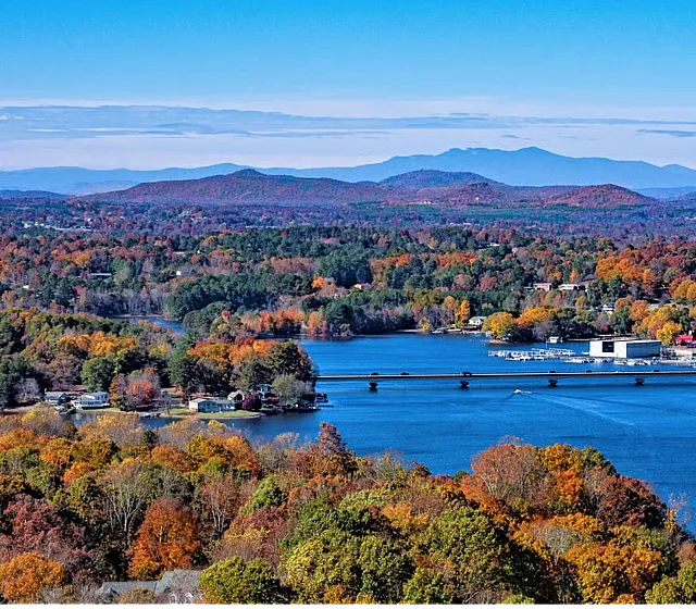 Breathtaking aerial view of a scenic lake surrounded by vibrant autumn foliage, with mountains in the background under a clear blue sky.