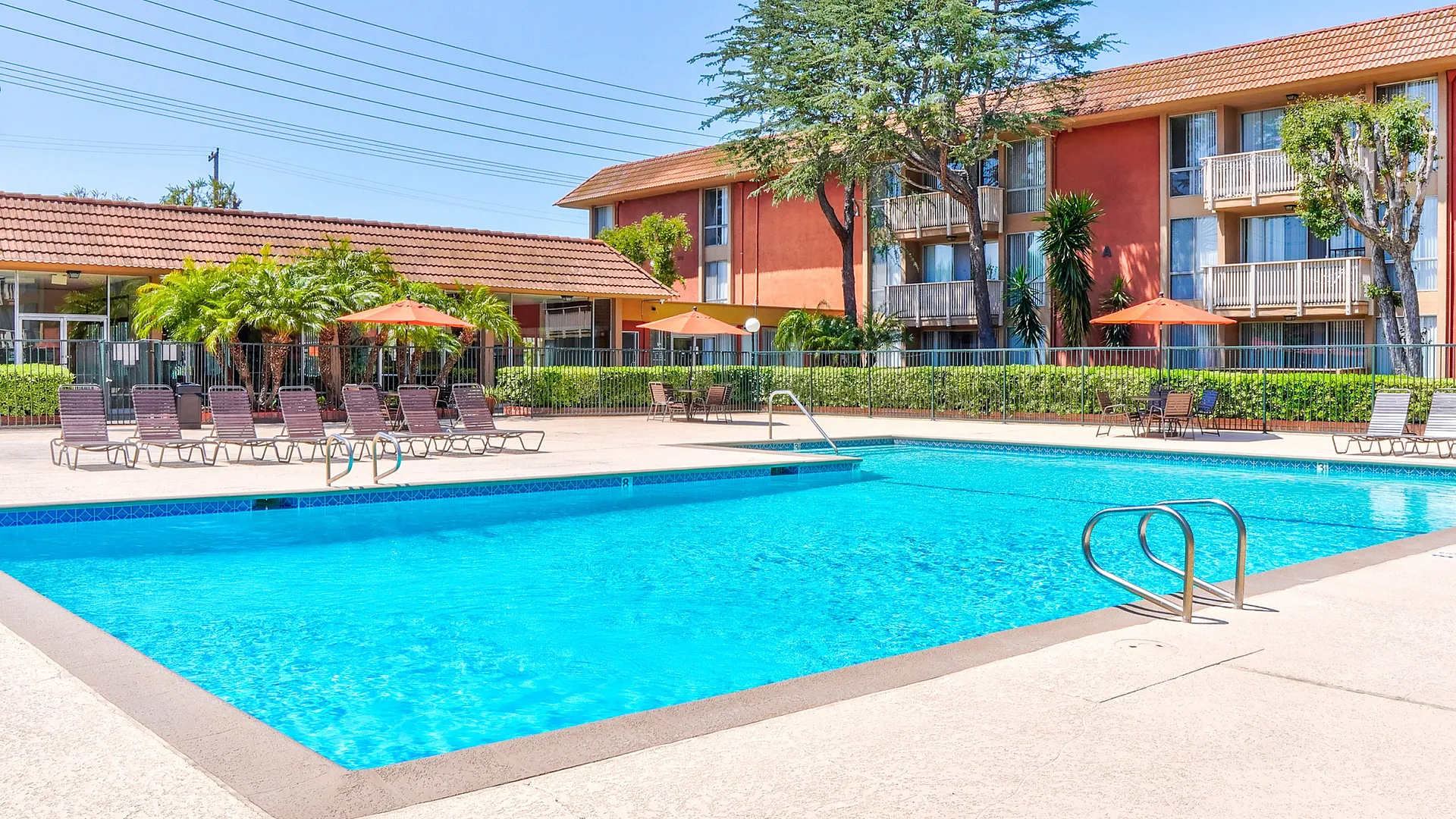 Inviting apartment complex featuring a sparkling swimming pool surrounded by sun loungers, tropical plants, and a beautiful red-brick building backdrop.