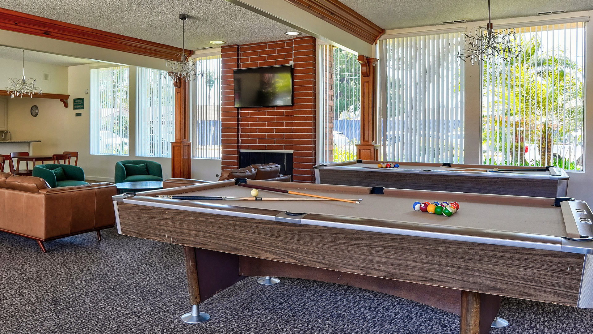 Inviting community lounge featuring pool tables, a cozy seating area, and a modern kitchen with a stylish brick fireplace and natural light, perfect for relaxation and entertainment.