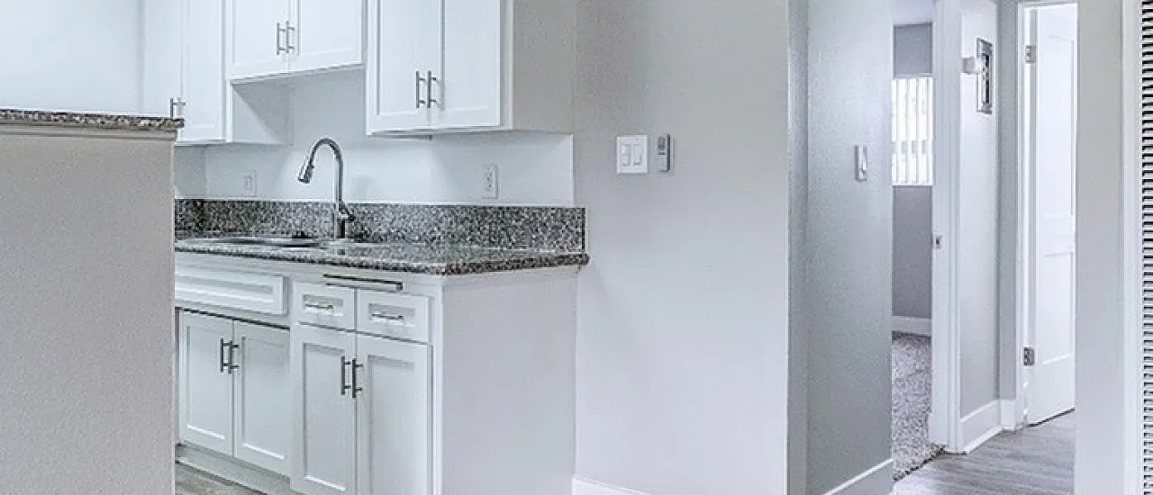 Modern apartment kitchen with sleek white cabinetry, granite countertops, and stylish ceiling fan, leading to a cozy hallway.
