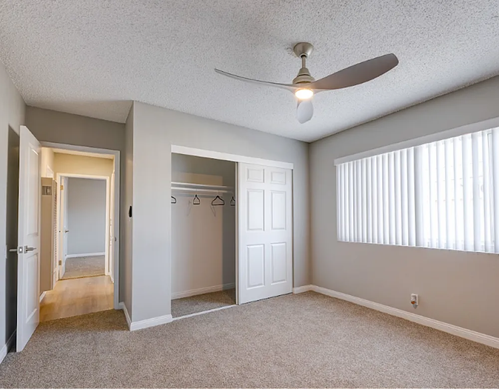 Bright and spacious bedroom featuring plush carpet, a ceiling fan, and large window with vertical blinds, perfect for a calming retreat.