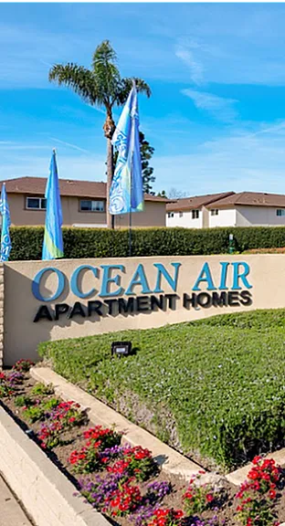 Inviting exterior of Ocean Air Apartment Homes with lush landscaping, palm trees, and vibrant signage showcasing 'Welcome Home' and 'Now Leasing'.