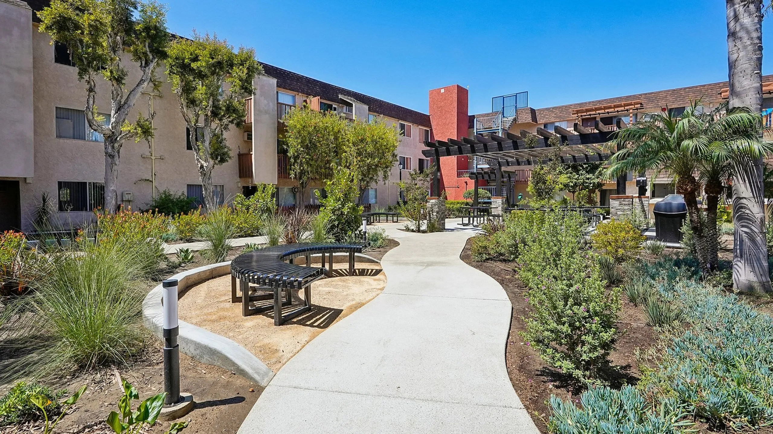Charming apartment complex with lush landscaping, walking paths, and outdoor seating areas under a clear blue sky.