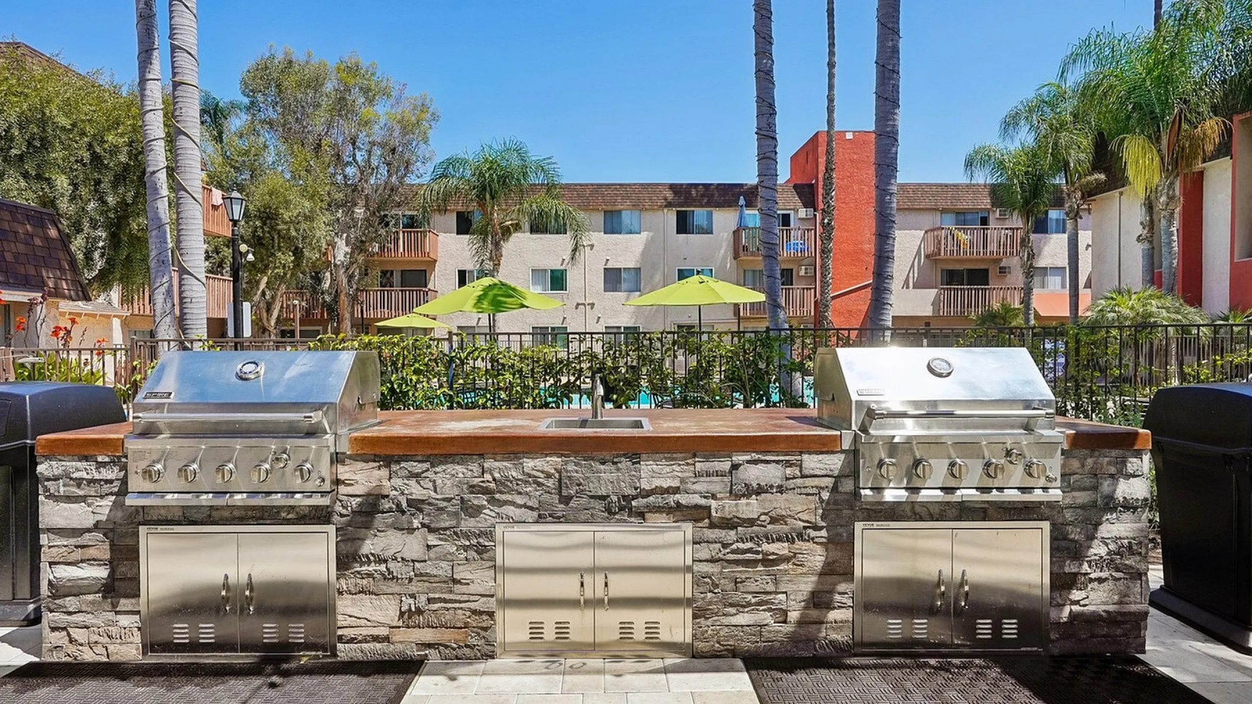 Luxurious outdoor BBQ area with stainless steel grills and lush palm trees, perfect for entertaining in a vibrant apartment complex setting.