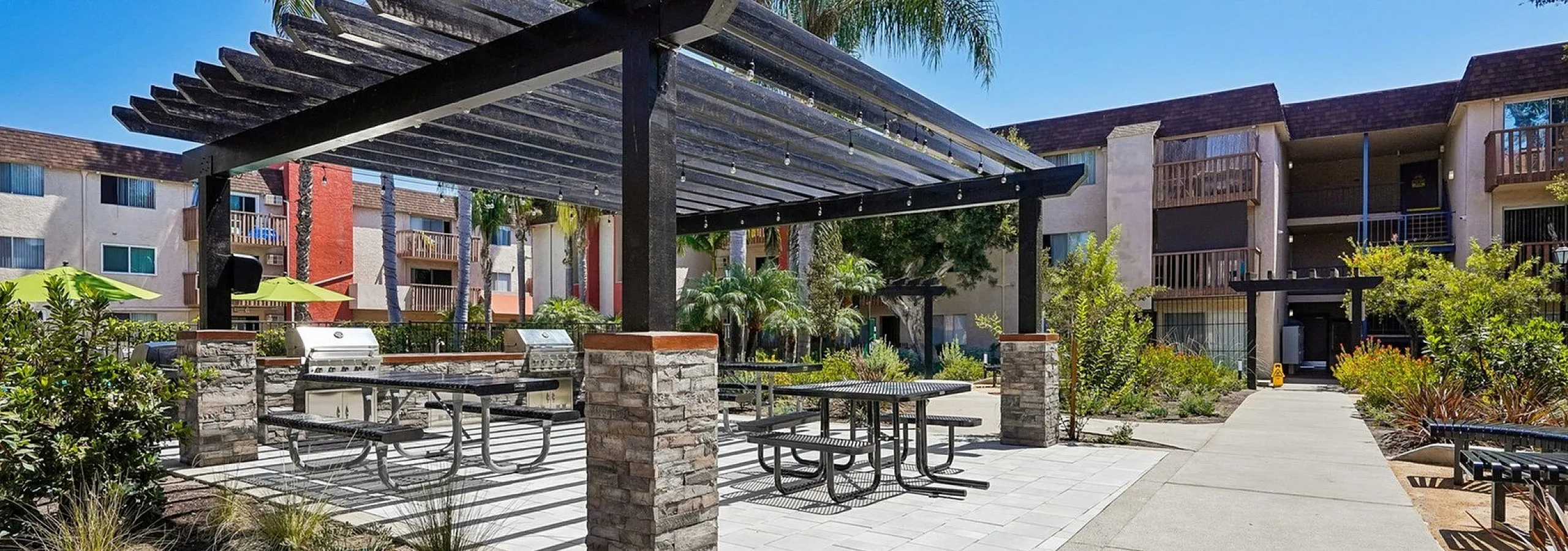 Inviting outdoor area with modern pergola, picnic tables, and grills, nestled amidst lush greenery in an upscale apartment complex.