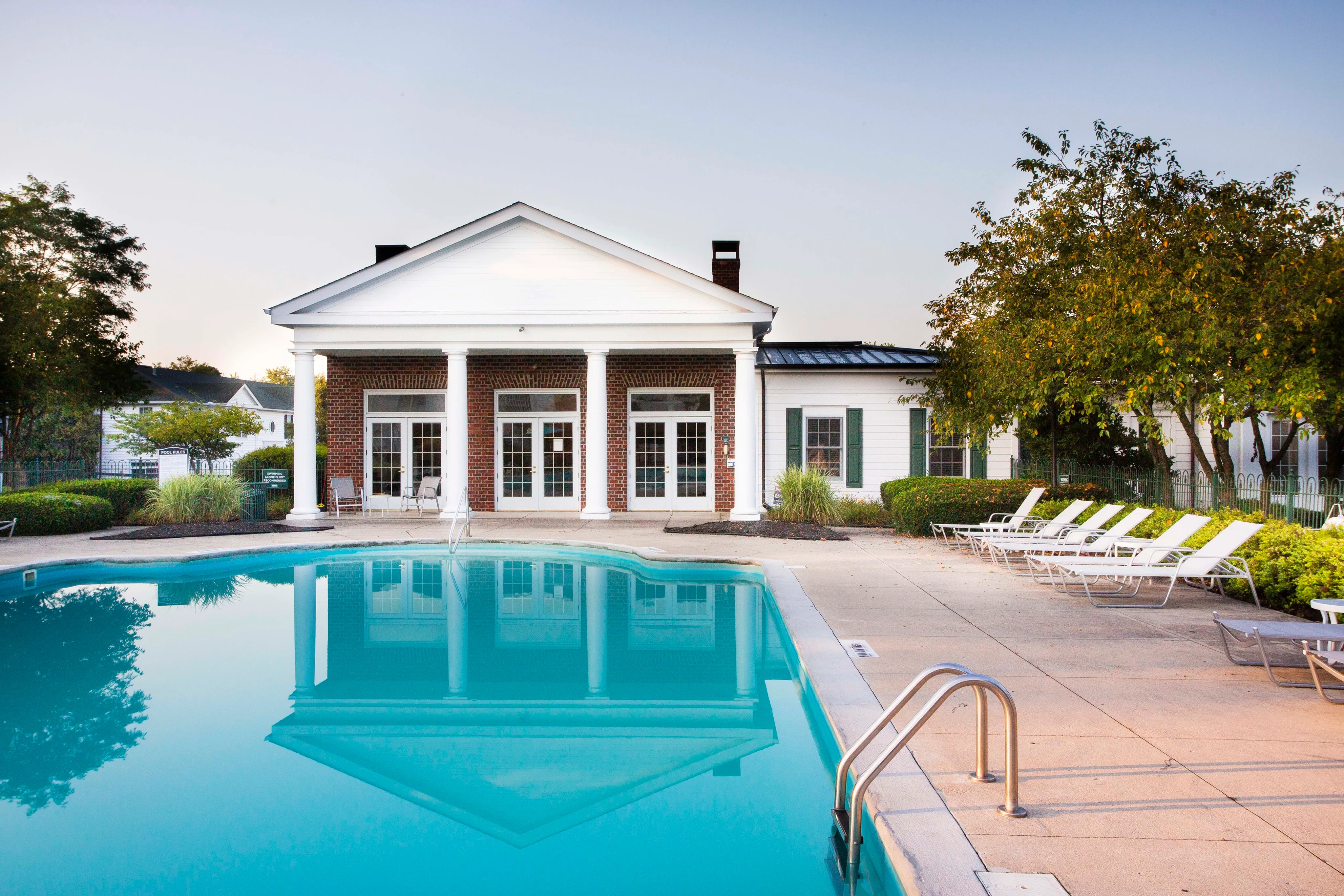 Charming poolside view of a modern apartment complex with elegant white columns and inviting lounge chairs, offering a perfect blend of relaxation and sophistication.