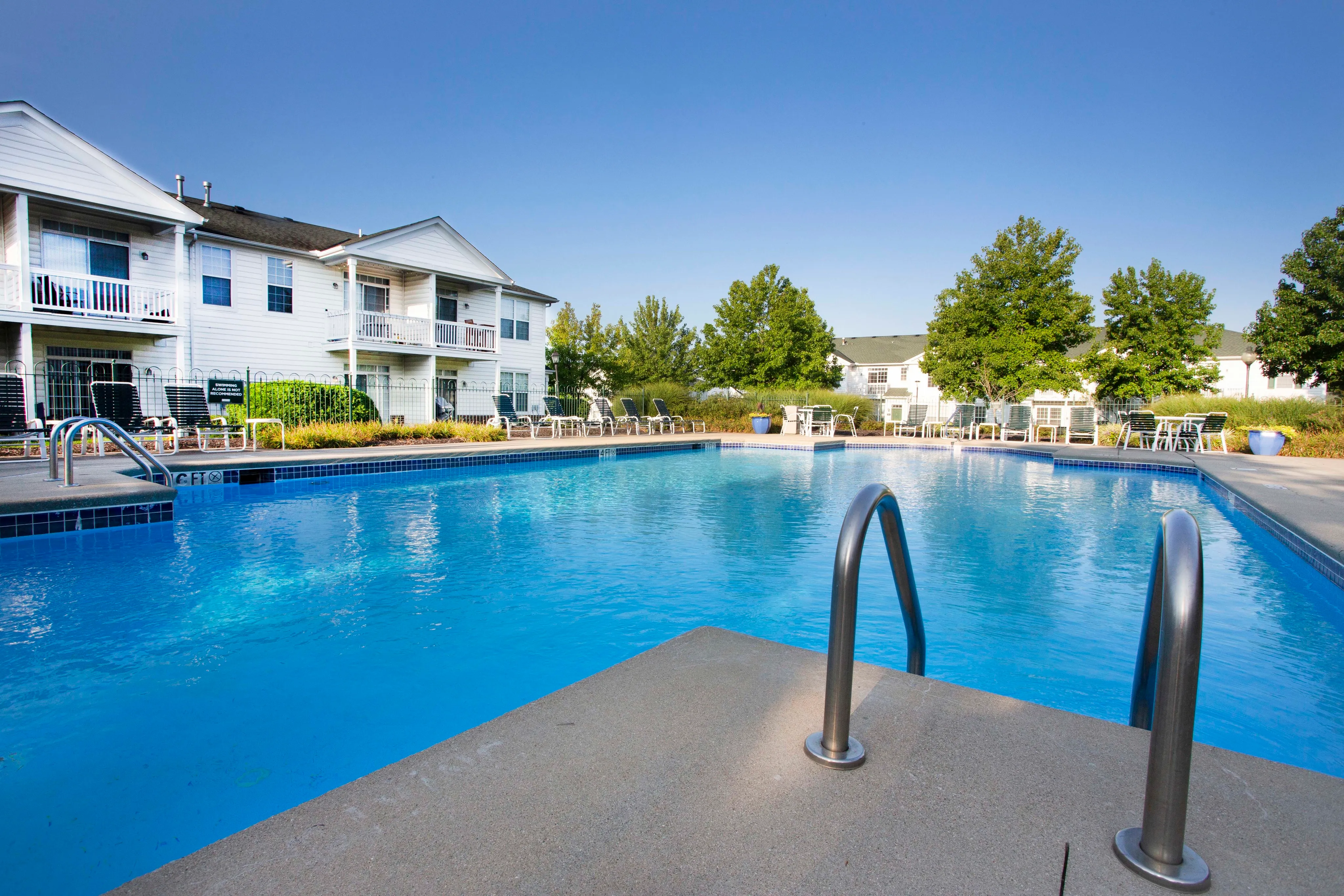 Inviting apartment complex with a sparkling outdoor swimming pool, surrounded by lush greenery and stylish lounge chairs, perfect for relaxing under the sun.