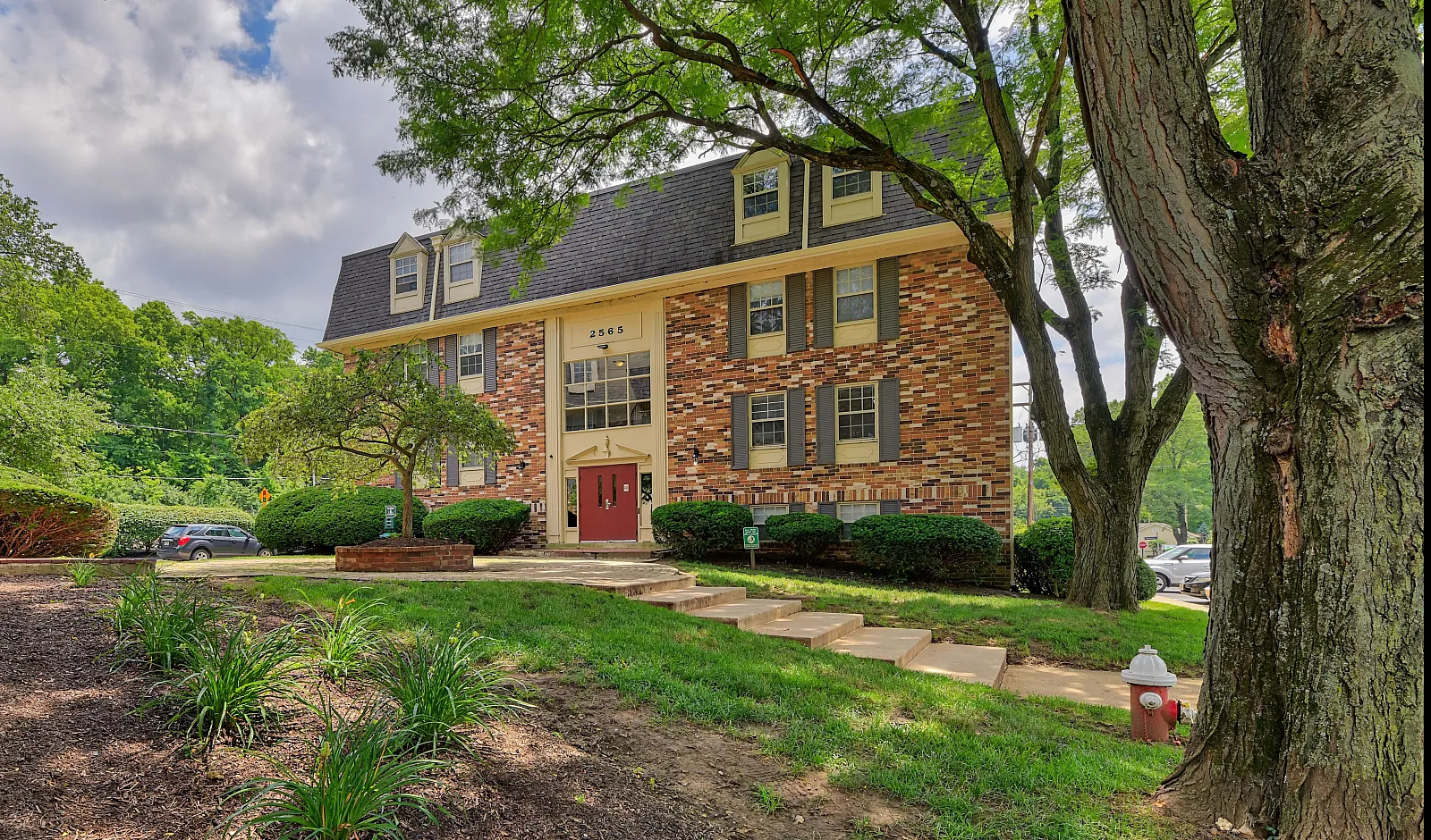 Charming brick apartment building with elegant red doors and lush landscaping, nestled in a serene, tree-lined environment.