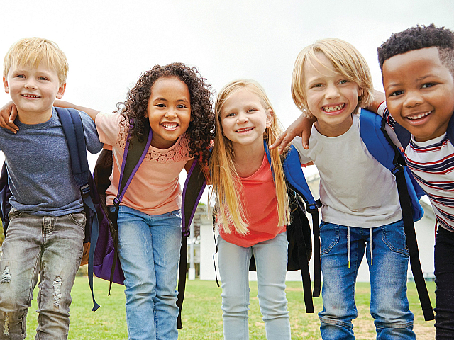 A group of smiling children with backpacks standing closely together in a green park.