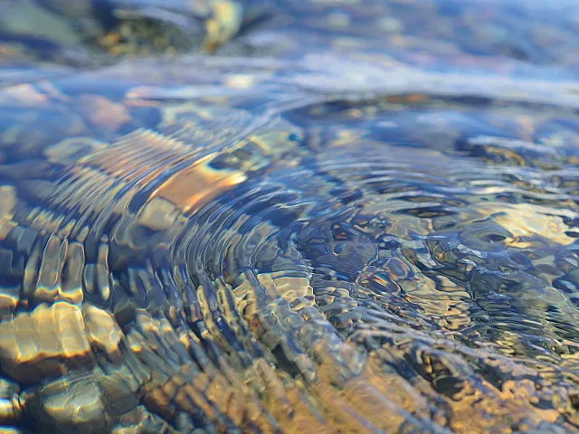 Rippling water with mesmerizing light reflections and colorful stones beneath, creating a tranquil and vibrant scene.