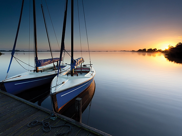 Peaceful lake at sunrise with two elegant sailboats docked, reflecting serene beauty and adventure.