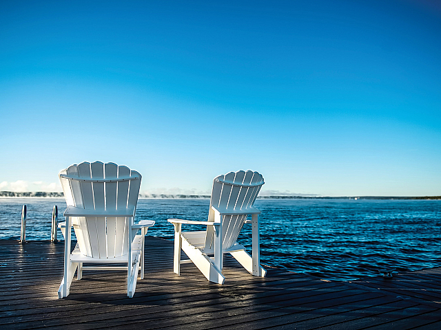 Tranquil lakeside view with two white Adirondack chairs on a wooden deck under a clear blue sky.