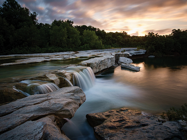 Tranquil natural landscape featuring a scenic waterfall cascading over smooth rocks, surrounded by lush greenery under a vibrant sunset sky.