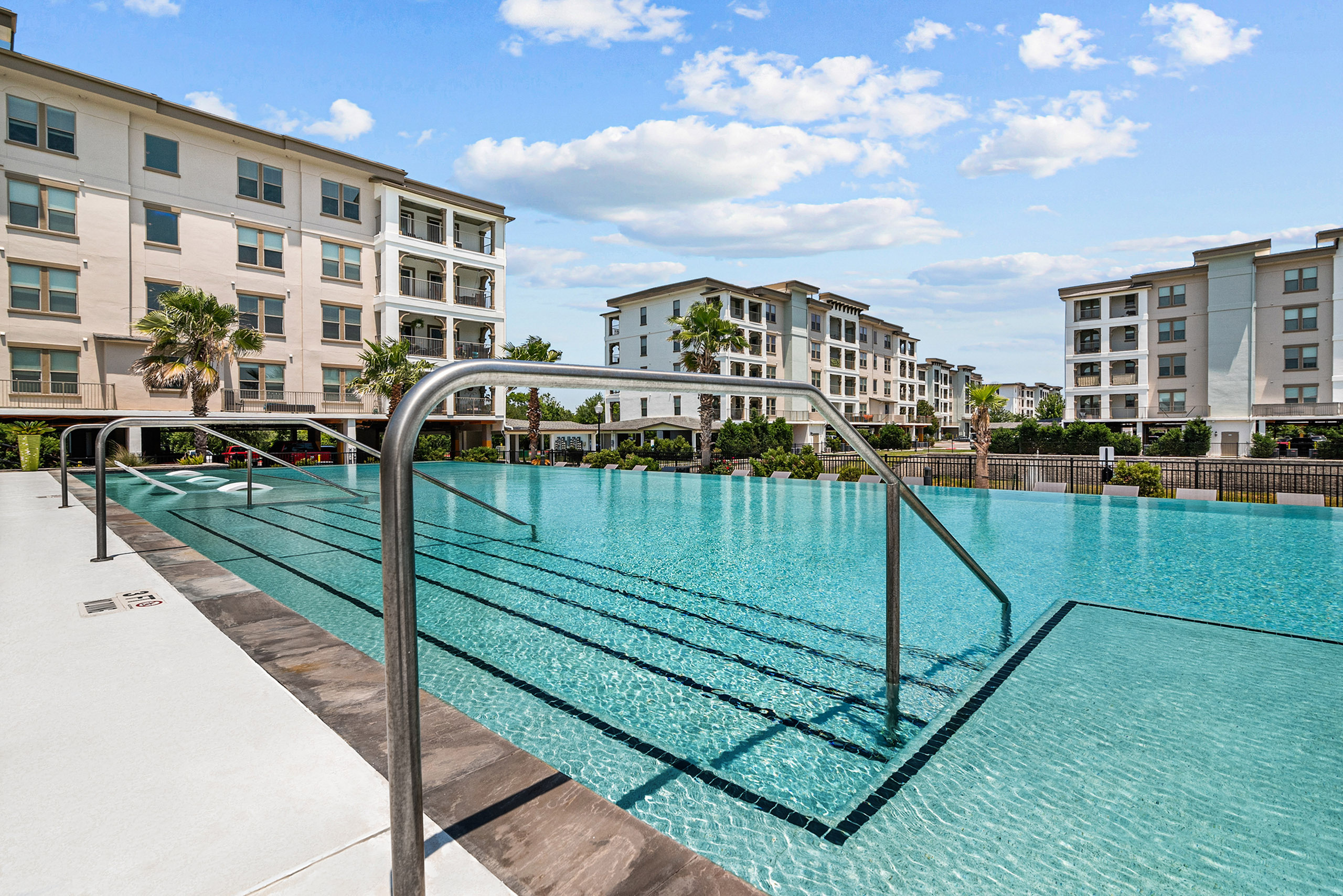 Luxurious infinity-edge apartment community with a sparkling swimming pool surrounded by modern buildings and lush greenery.